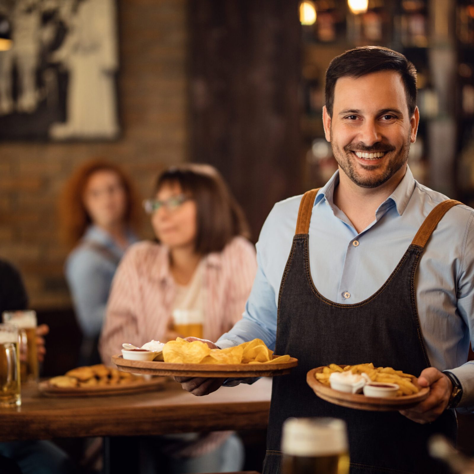 Happy waiter holding plates with food and looking at camera while serving guests in a restaurant.